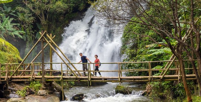 Eine Frau und ein Mann laufen Hand in Hand über eine Holzbrücke über einem Fluss, im Hintergrund ist ein großer Wasserfall