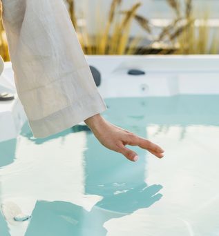 Eine Frau sitzt im Bademantel am Rand eines Whirlpools und hält Ihre Hand über die Wasseroberfläche
