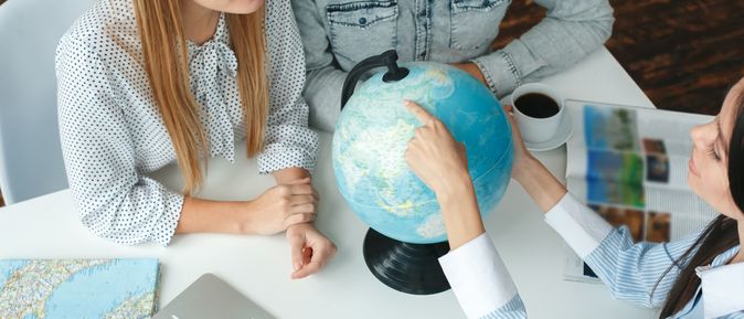 Ein Paar sitzt mit einer Reiseberaterin am Tisch, während die Reiseberaterin dem Paar etwas auf einem Globus zeigt