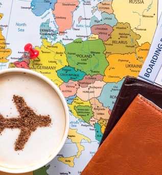 Landkarte mit einem Pin, der in Deutschland gesteckt ist. Auf der Karte steht eine Tasse Kaffee, wo der Schaum die Form einen Flugzeugs ausspart. Ein Flugticket und Lederbörse liegen auch auf der Karte