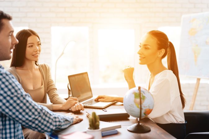 Ein Paar sitzt mit einer Reiseberaterin am Tisch, während das Licht durch das Fenster im Hintergrund den Raum erleuchtet