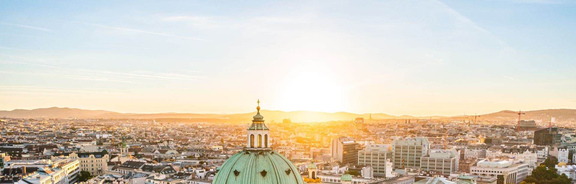 Sonnenaufgang über einer Stadt. Im Vordergrund ist ein weißes Bauwerk mit grünen Kuppeln