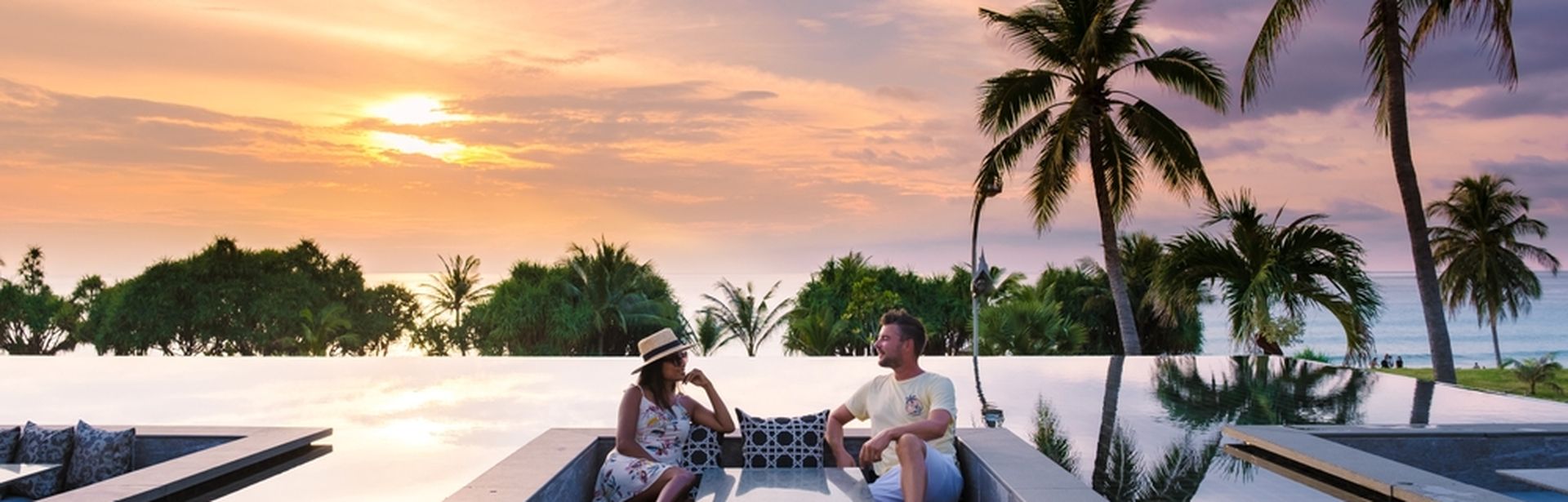 Paar sitzt in einer versenkten Lounge im Infinity-Pool bei Sonnenuntergang, umgeben von Palmen und Meerblick