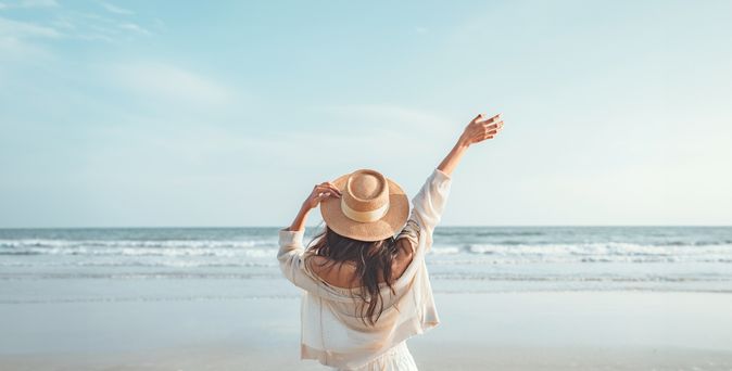 Eine Frau mit Hut von hinten, Sie schaut vom Strand aus auf das Meer und hebt den Arm 