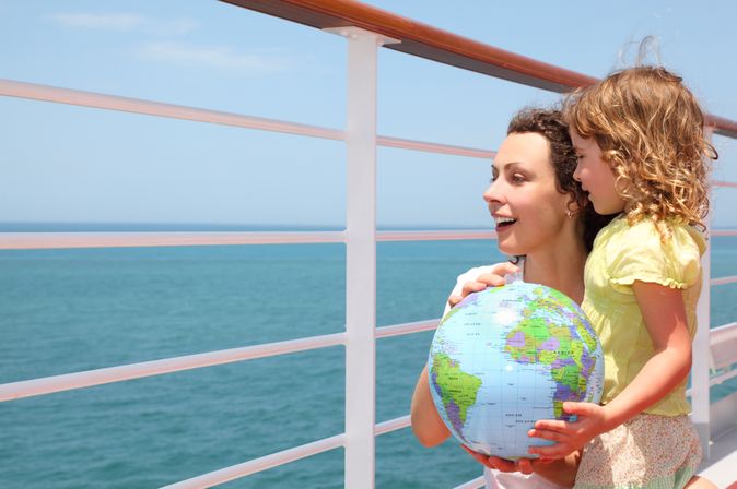 Mutter und Kind schauen über das Geländer eines Kreuzfahrtschiffes und halten einen Globus