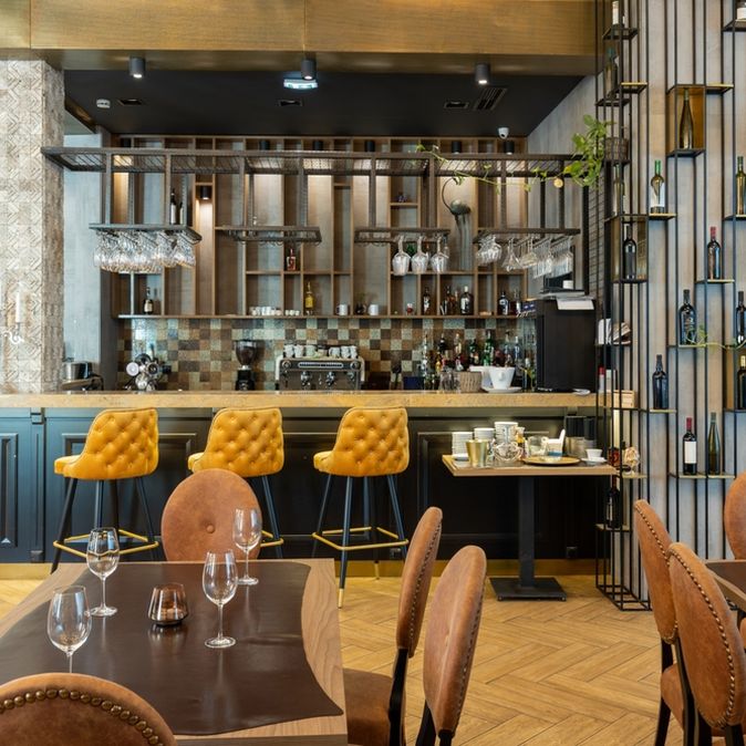 Eine Modern eingerichtete Bar mit gelben Barhockern und einer Wand mit Weinflaschen, im Vordergrund stehen mehrere mit Gläsern gedeckte Tische