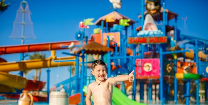 Ein Junge in einer gelben Badehosen steht vor einem großen Aquapark und zeigt einen Daumen nach oben
