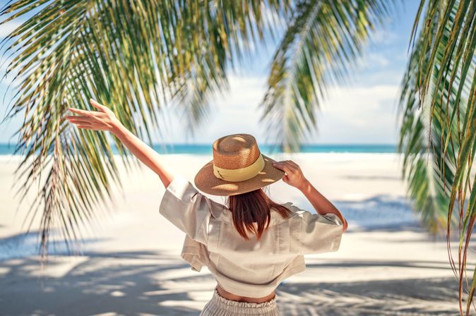 Frau mit Sonnenhut am Strand umgeben von Palmenblättern hebt einen Arm in die Luft