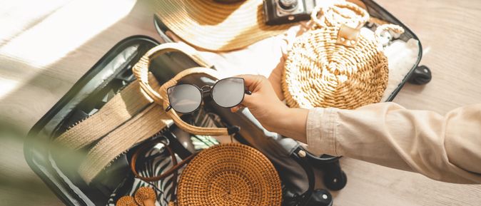 Ein offener Koffer, eine Hand legt eine Sonnenbrille zu den bereits gepackten Dingen