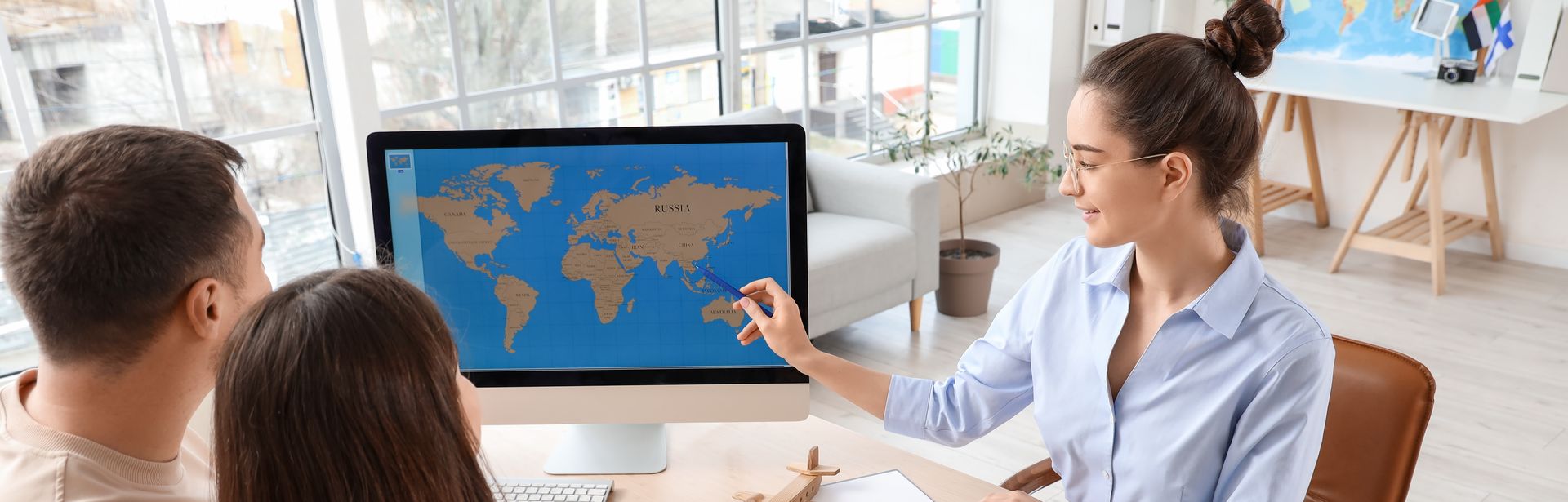 Ein Paar sitzt mit einer Reiseberaterin am Tisch. Die Reiseberaterin zeigt auf eine Weltkarte auf einem Bildschirm. Der Mann hat den Arm um seine Freundin gelegt