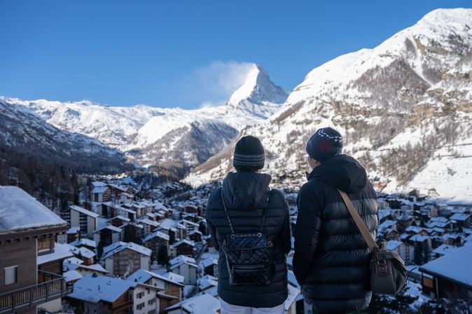 Zwei Personen stehen erhöht und schauen auf ein verschneites Dorf in den Bergen
