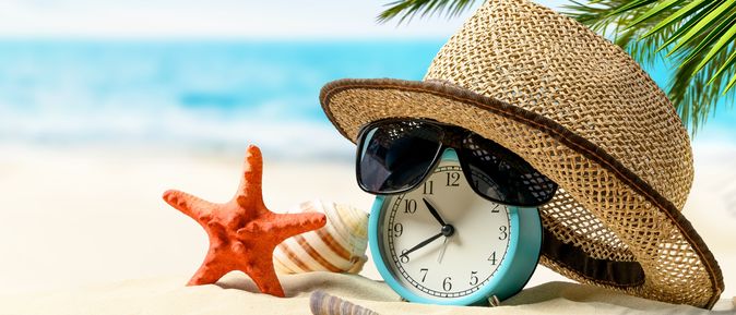 Ein Wecker mit Sonnenhut und einer Sonnenbrille liegen am Strand; Daneben befinden sich Muscheln und ein Seestern