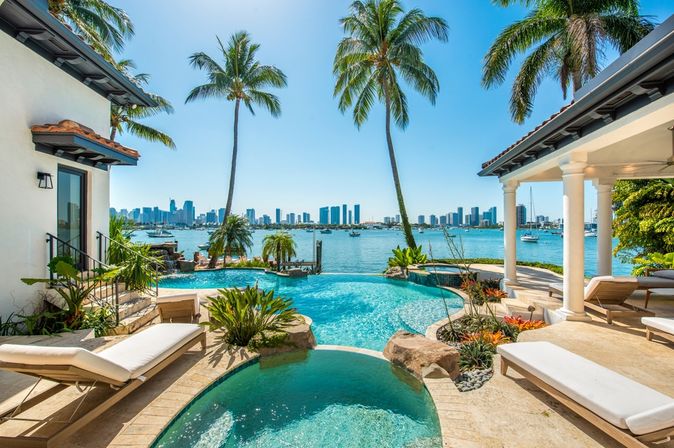 Von einem Pool umgeben von Palmen schaut man auf das Meer, im Hintergrund Hochhäuser