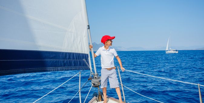 Junge mit roter Kappe steht vorne auf einem Segelboot und schaut auf das Meer