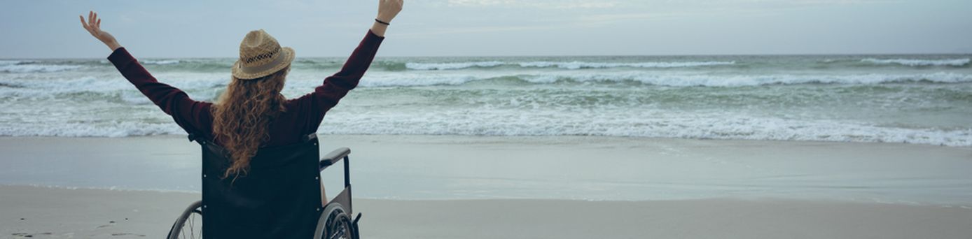 Frau im Rollstuhl hebt die Arme und schaut aufs Meer