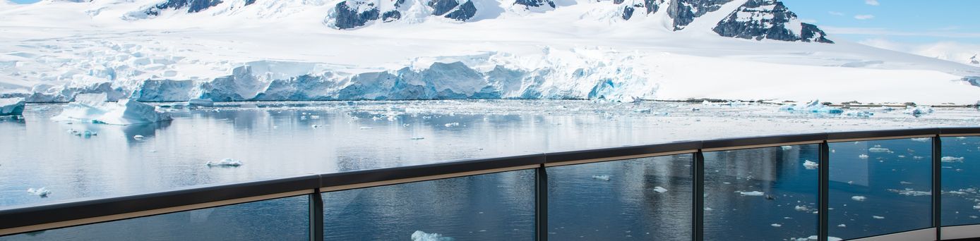 Ein Schiffsdeck mit Glasgeländer bietet einen Blick auf eine antarktische Landschaft mit schneebedeckten Bergen und schwimmenden Eisschollen
