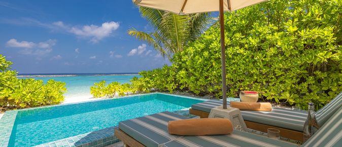 Zwei Liegen unter einem Sonnenschirm, davor ein kleiner Pool und der Strand 