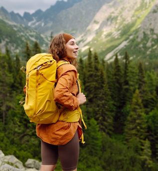 Eine Frau mit gelbem Wanderrucksack steht auf einem Berg und schaut begeistert in die Ferne