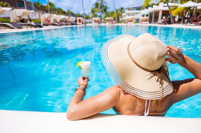 Eine Frau mit Hut von hinten, sie sitzt in einem Pool und hat ein weißes Getränk in der Hand