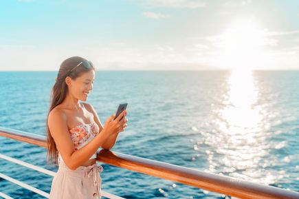 Eine Frau steht lächelnd an der Reling eines Kreuzfahrtschiffes und schaut auf ihr Smartphone