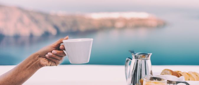 Ein gedeckter Tisch mit einer Kanne und Tellern mit Obst, eine Hand hält eine Tasse hoch, im Hintergrund ist das Meer