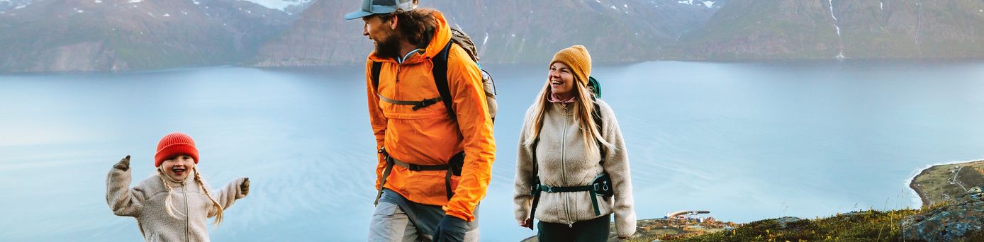 Eine Familie wandert in Winterkleidung an einem Gewässer, im Hintergrund sind verschwommen Berge 