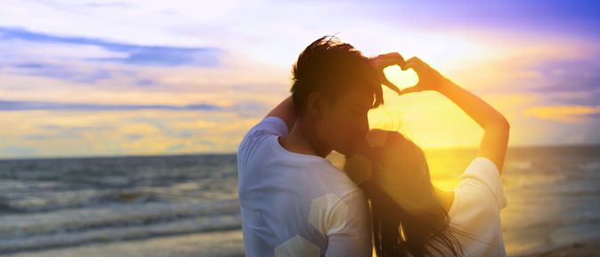 Ein Paar formt mit den Händen gemeinsam ein Herz und schaut bei Sonnenuntergang auf das Meer