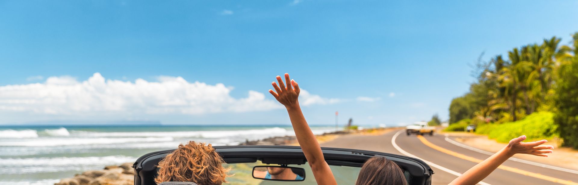 Zwei Personen im Auto mit offenem Verdeck, im Hintergrund kann man das Meer sehen