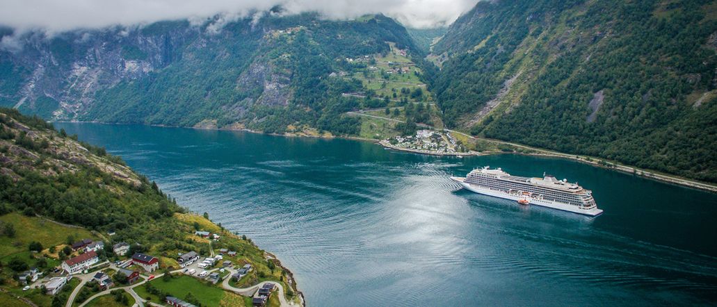 Ein Kreuzfahrtschiff fährt durch einen tiefen, von grünen Bergen gesäumten Fjord mit einem kleinen Dorf im Vordergrund.