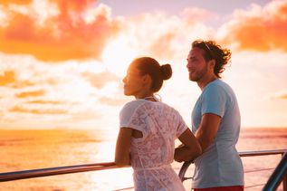 Eine Frau und ein Mann stehen bei Sonnenuntergang an der Reling eines Kreuzfahrtschiffes