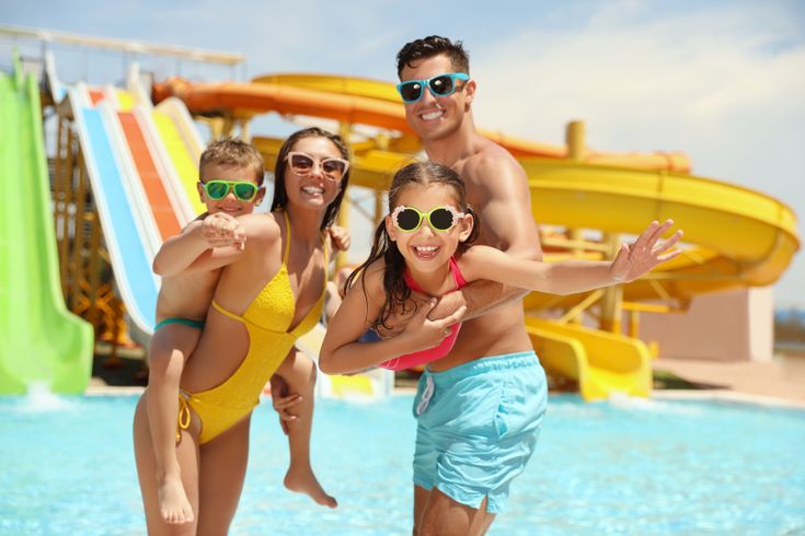 Eine Familie steht in einem Pool vor einigen Wasserrutschen, die Frau hat eins der Kinder auf dem Rücken, der Mann trägt das zweite Kind im Arm 