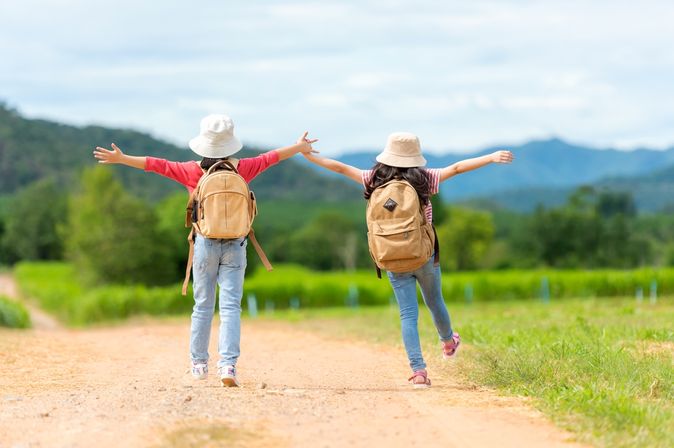 Zwei Mädchen mit Rucksäcken laufen fröhlich einen Weg entlang und heben die Arme in die Luft