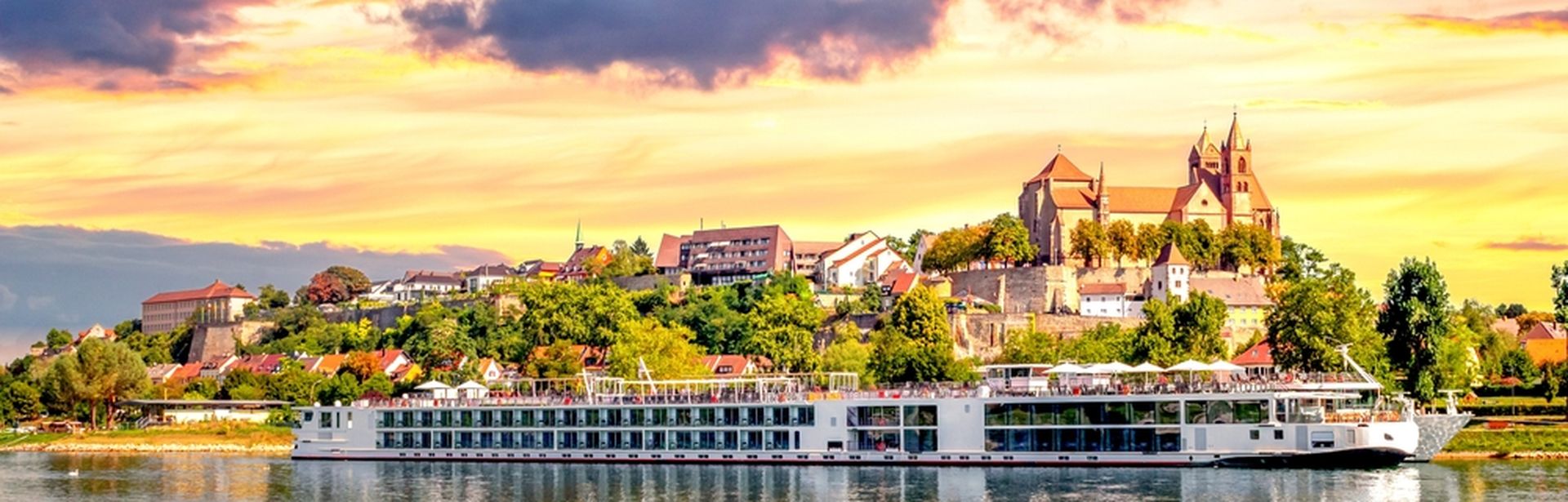 Langes Flusskreuzfahrschiff fährt entlang eines kleinen Dorfes, bei gelb leuchtendem Himmel