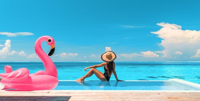 Frau sitzt am Rand eines Pools mit Meerblick, neben Ihr ein aufblasbarer Flamingo