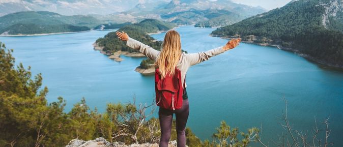 Eine Frau trägt einen roten Rucksack, streckt die Arme aus und schaut von einem Berg runter auf einen See mit Insel