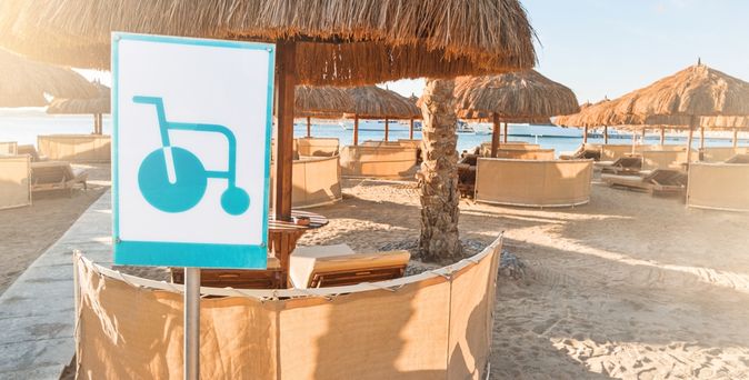 Strand mit mehreren Tischen und Schirmen, ein Schild im Vordergrund auf dem ein Rollstuhl abgebildet ist