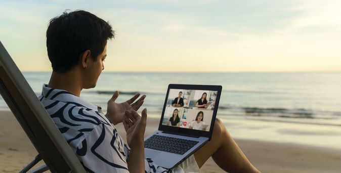 Ein Mann sitzt in einem Liegestuhl am Meer mit Laptop auf dem Schoß und nimmt an einer Videokonferenz teil