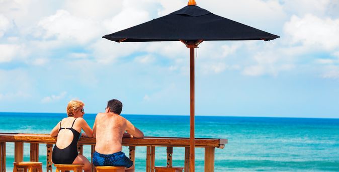 Eine Frau und ein Mann in Badekleidung sitzen an einer Holztheke auf Holzstühlen vor einem Pool und schauen auf das Meer