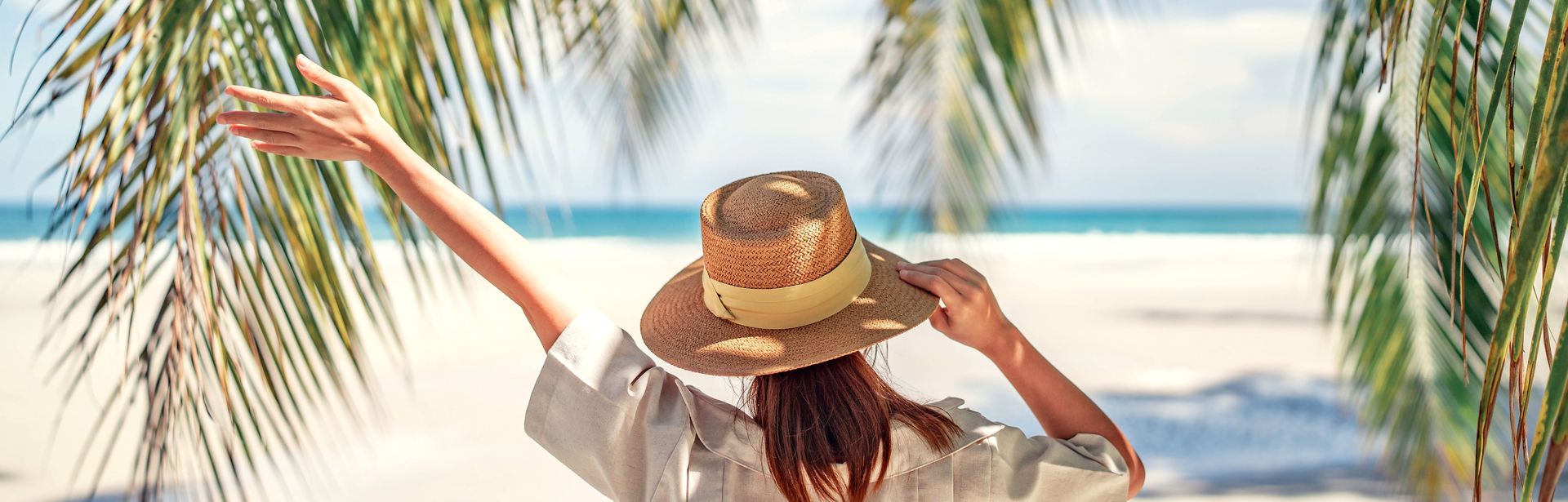 Frau mit Sonnenhut am Strand umgeben von Palmenblättern hebt einen Arm in die Luft