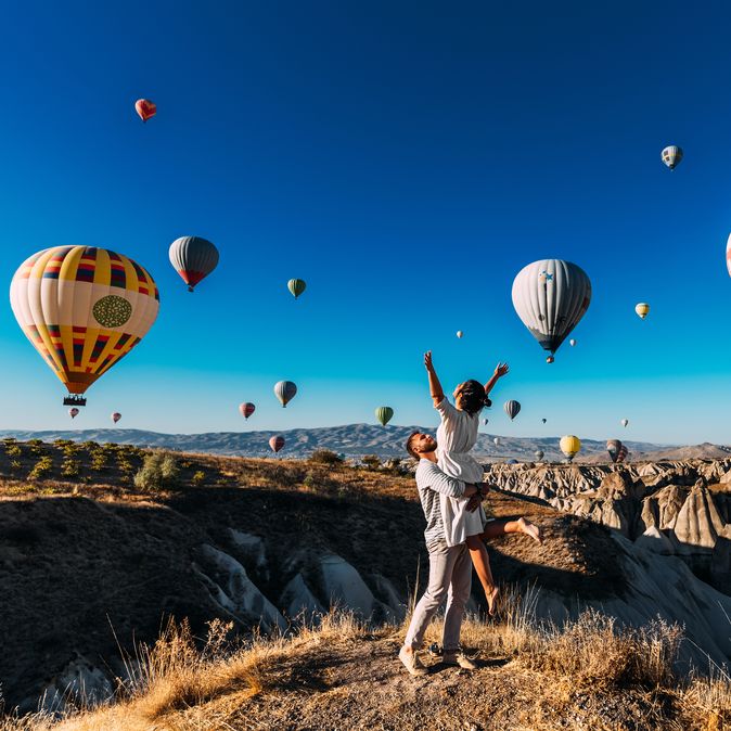 Ein Mann trägt eine Frau im Arm, um Sie herum schweben Heißluftballons durch die Luft
