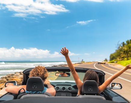 Zwei Personen fahren in einem Auto eine Straße am Meer entlang, eine von ihnen reißt die Arme nach oben