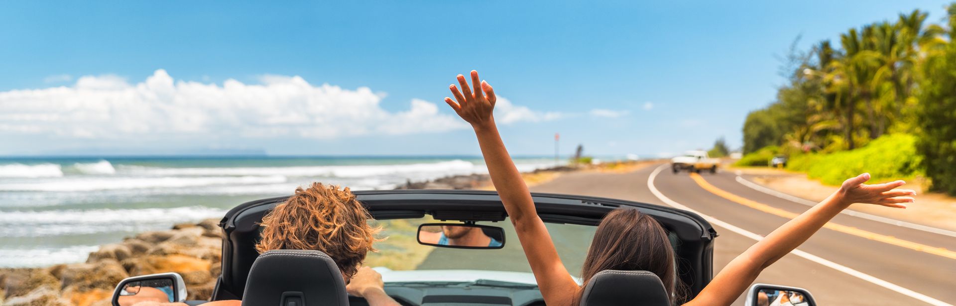 Zwei Personen im Auto mit offenem Verdeck, im Hintergrund kann man das Meer sehen