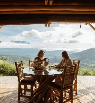 Auf einer Terrasse mit beeindruckendem Ausblick auf eine grüne Landschaft sitzen zwei Personen an einem Tisch 