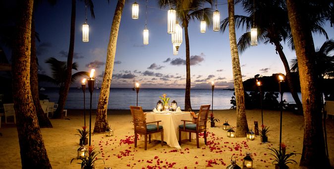 Ein gedeckter Tisch am Strand, Rosenblätter liegen am Boden und Laternen hängen im Baum