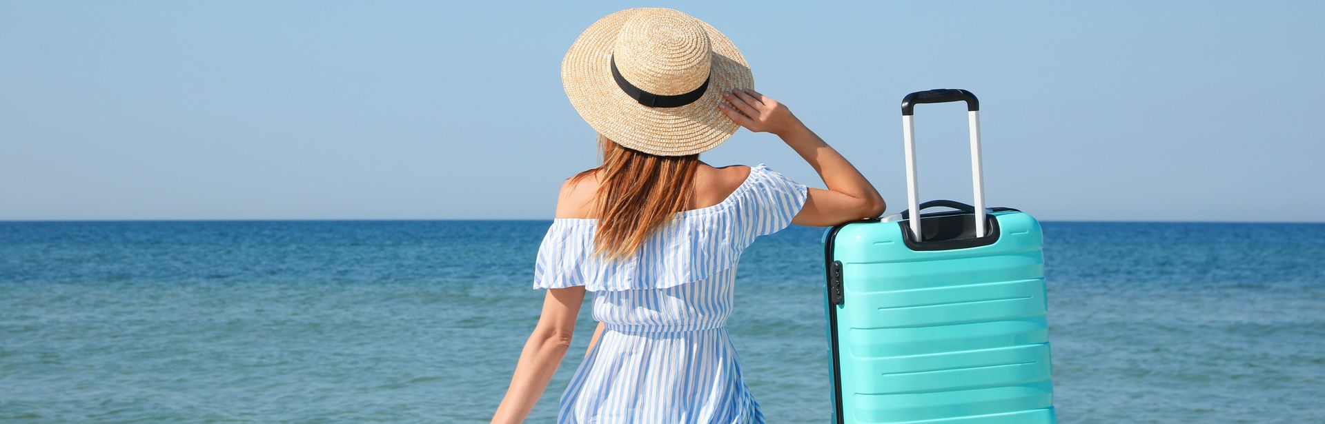 Frau im Sommerkleid und Sonnenhut sitzt am Meer und schaut auf das Meer. Ihr rechter Arm ist auf einen blauen Koffer gelehnt