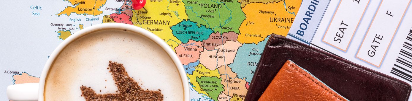 Landkarte mit einem Pin, der in Deutschland gesteckt ist. Auf der Karte steht eine Tasse Kaffee, wo der Schaum die Form einen Flugzeugs ausspart. Ein Flugticket und Lederbörse liegen auch auf der Karte