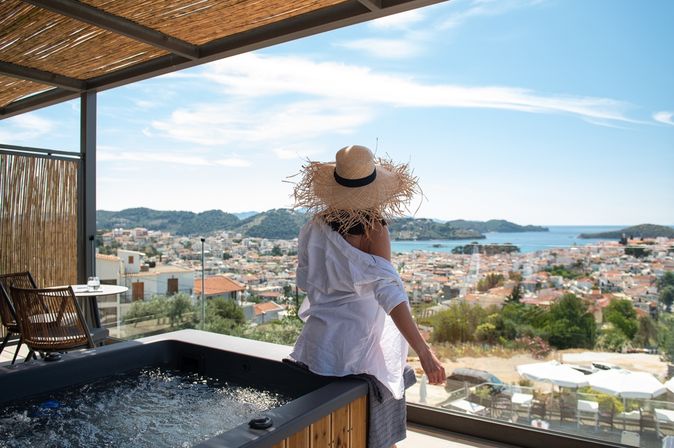 Eine Frau mit Sonnenhut sitzt auf dem Rand eines Whirlpools auf einem Balkon und blickt runter Richtung der Stadt