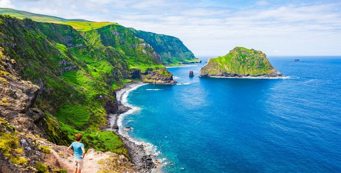 Ein Junge steht erhöht auf einem Hügel, vor ihm das Meer. Im Hintergrund sieht man grüne Wiesen und eine kleine Insel im Meer