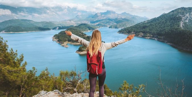 Eine Frau mit Rucksack schaut von einer Klippe auf einen großen See, eine Berglandschaft und breitet die Arme aus