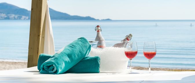 Zwei aufgerollte Handtücher liegen neben zwei Gläsern und ein Getränkekühler mit Eis und zwei Flaschen auf einer Decke am Strand